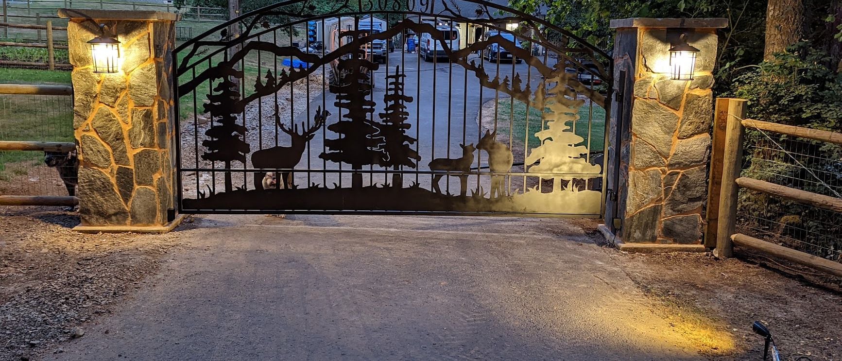 Lit stone entry columns and landscape lighting on a wooded property at dusk
